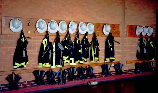 Firefighting gear including helmets, jackets, and boots are neatly hung on a brick wall.