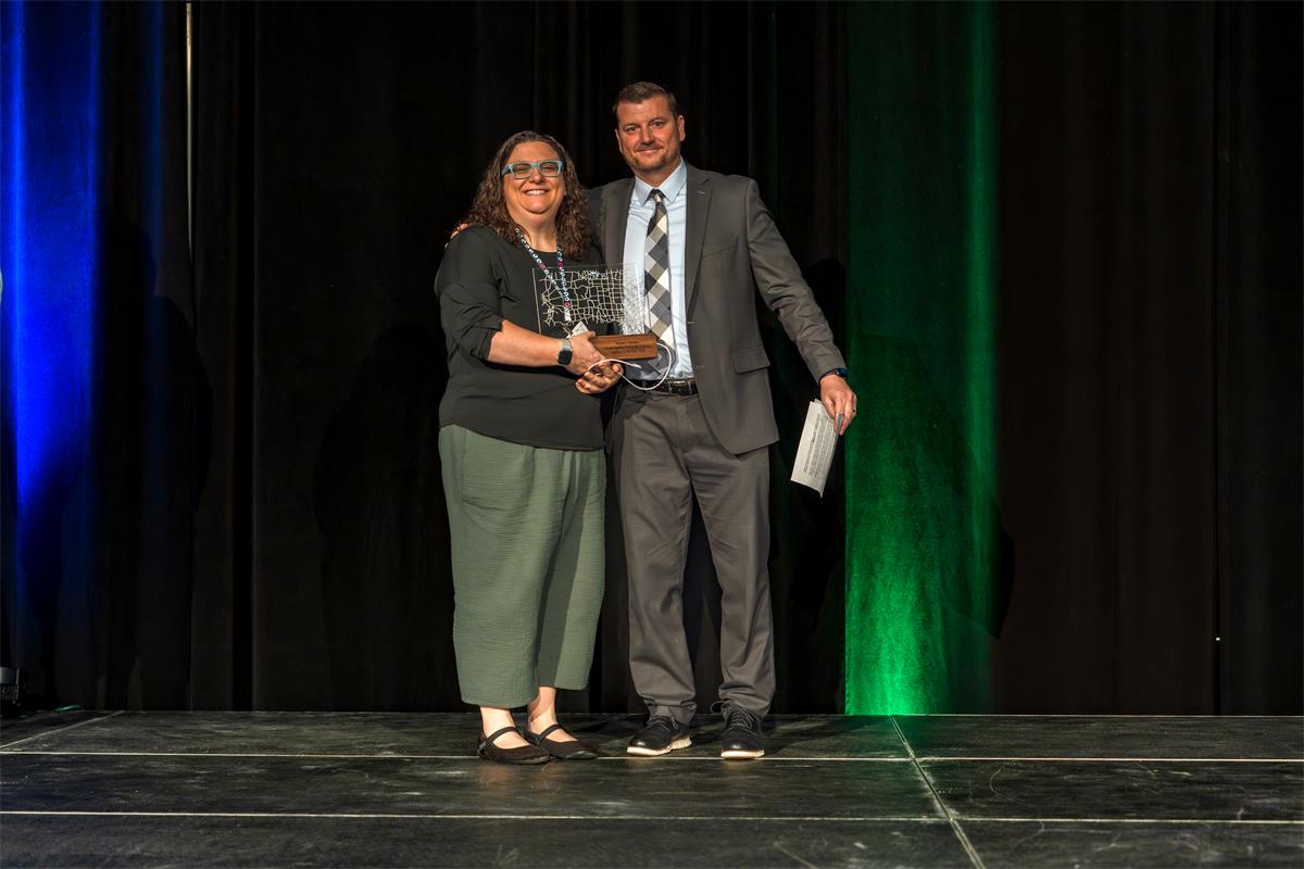 Rachel J. Pierson and Josh Siegfried stand together on stage, both smiling and holding a glass award on a wooden base.