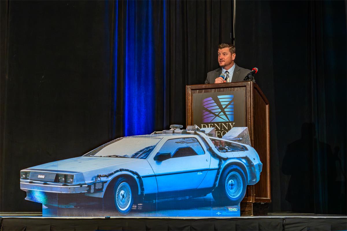 Assistant Principal Josh Siegfried speaks at a podium at TIE26, announcing the South Dakota Computer Science Teacher of the Year Award. A DeLorean cutout is visible on stage in front of the podium.