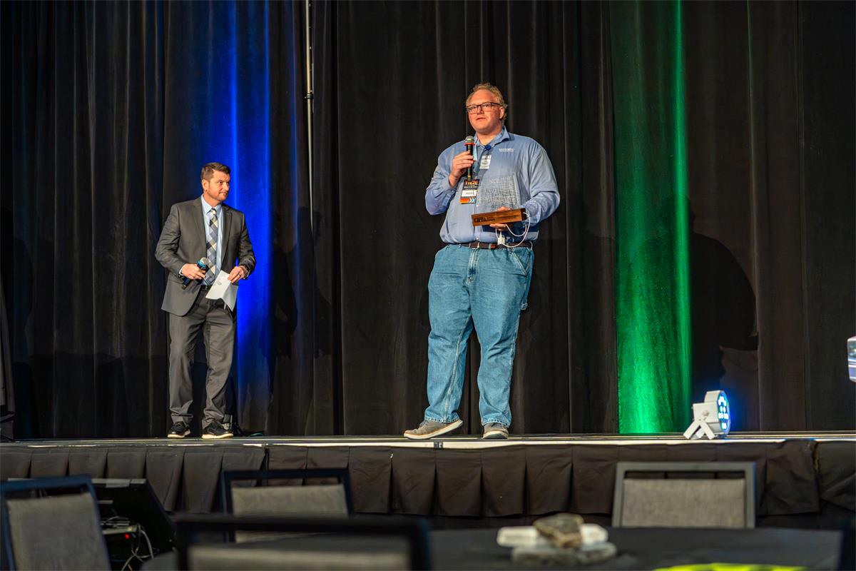 CSTA-SD chapter president Ben Benson speaks on stage at TIE26 holding the South Dakota Computer Science Teacher of the Year Award. Josh Siegfried, Assistant Principal at Memorial Middle School, looks on at left.