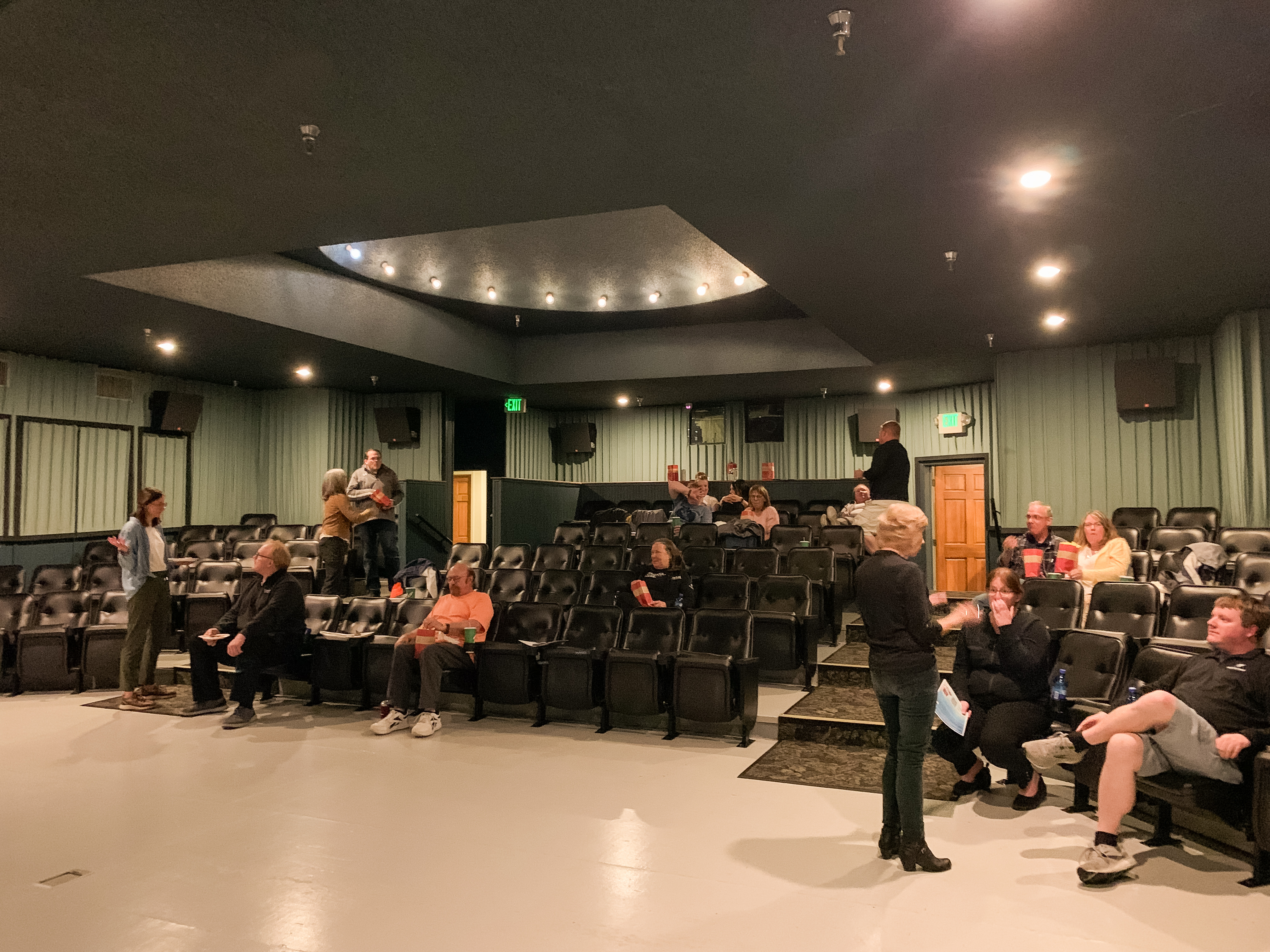 A small movie theater with green walls and black cushioned seats, where people are seated or standing and chatting before a presentation or event begins. The room is lit with overhead lights, and there is a person speaking near the front to a small group.