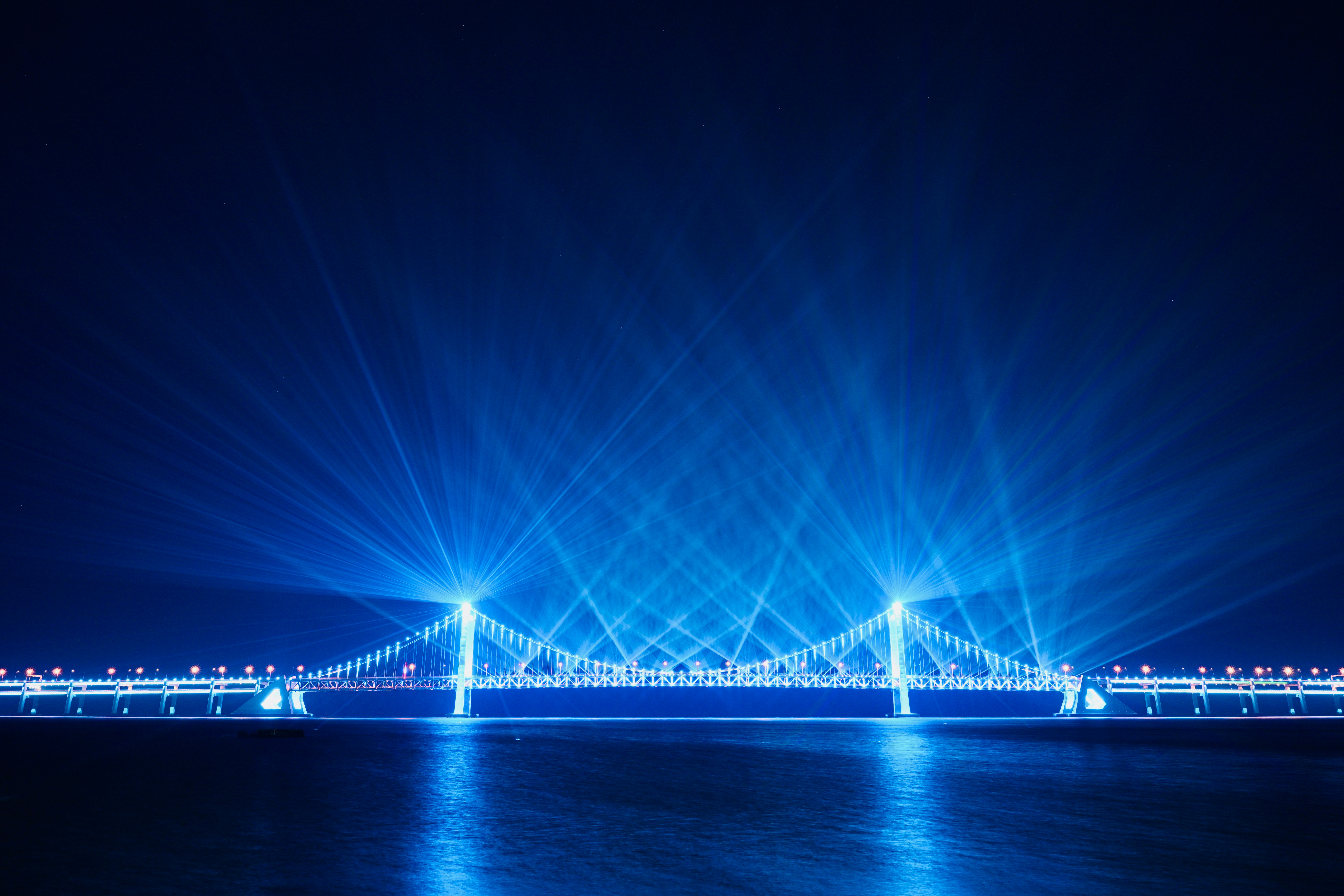 Illuminated suspension bridge at night glowing in blue with radiating light beams reflecting across calm water.