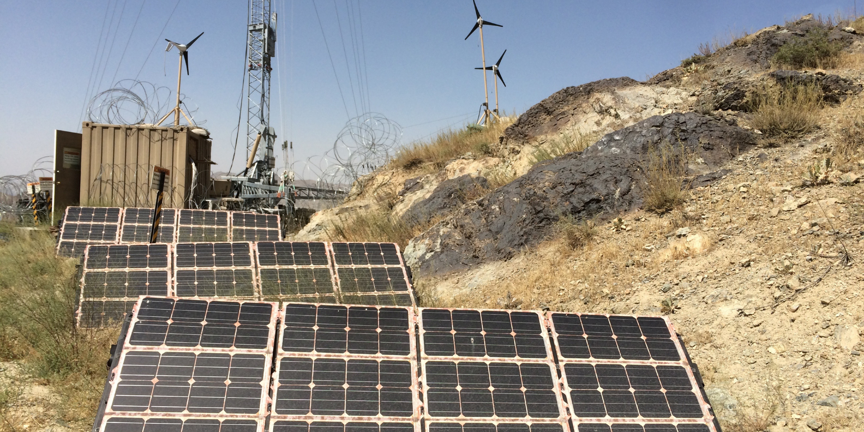 Solar panels in a desert with wind turbines in the background.