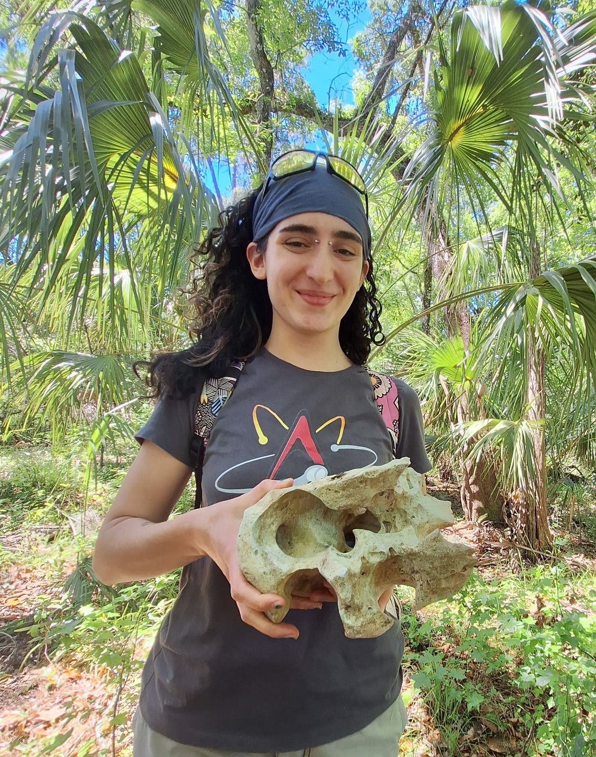 Smiling woman holding large animal skull; palm trees in background.
