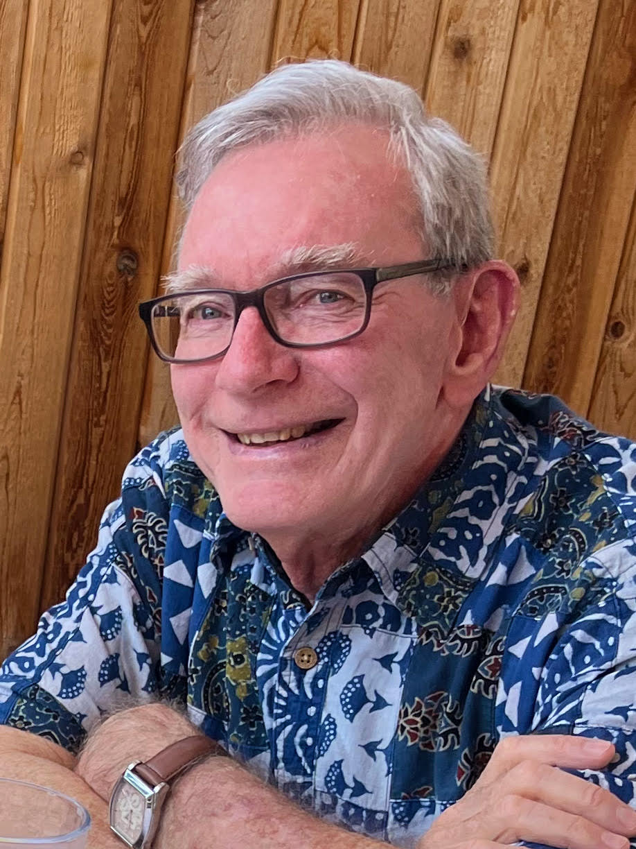A gentleman with black frame glasses who is wearing a colorful blue button up shirt smiles for the camera in front of a wooden fence.