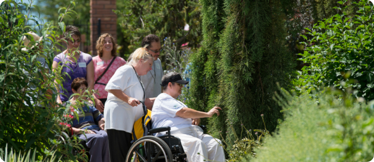 An individual in a wheelchair reaches out to touch greenery in a lush garden while people walk behind them