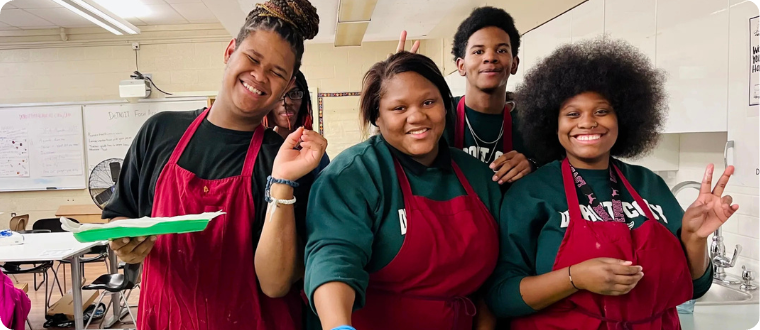 Four people with red aprons smile in a lunch line in a cafeteria