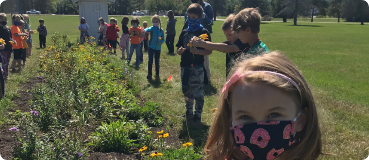 Kids are lined up on both sides of a long narrow flower bed, some holding flowers