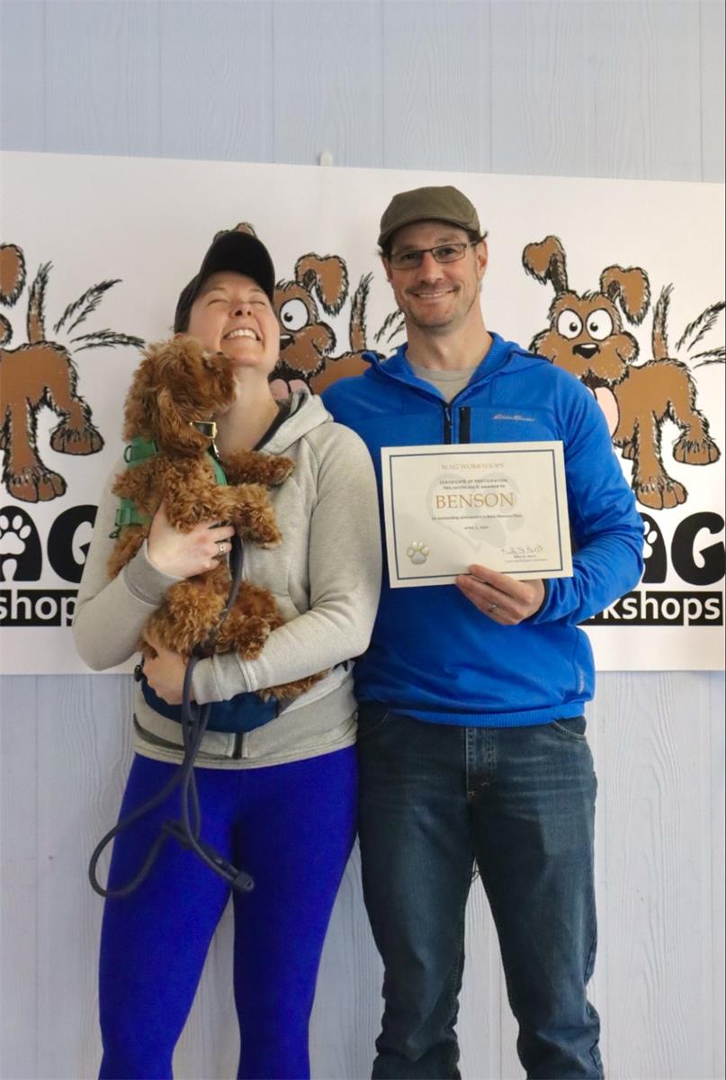 Woman and man hold their graduation certificate and a small dog in front of a Wag Workshops banne.