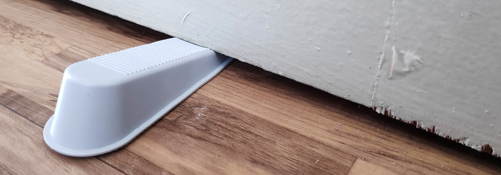 A white ribbed plastic doorstop holds a wooden door slightly open on a hardwood floor. The door’s edge shows visible wear, including scratches and dents.