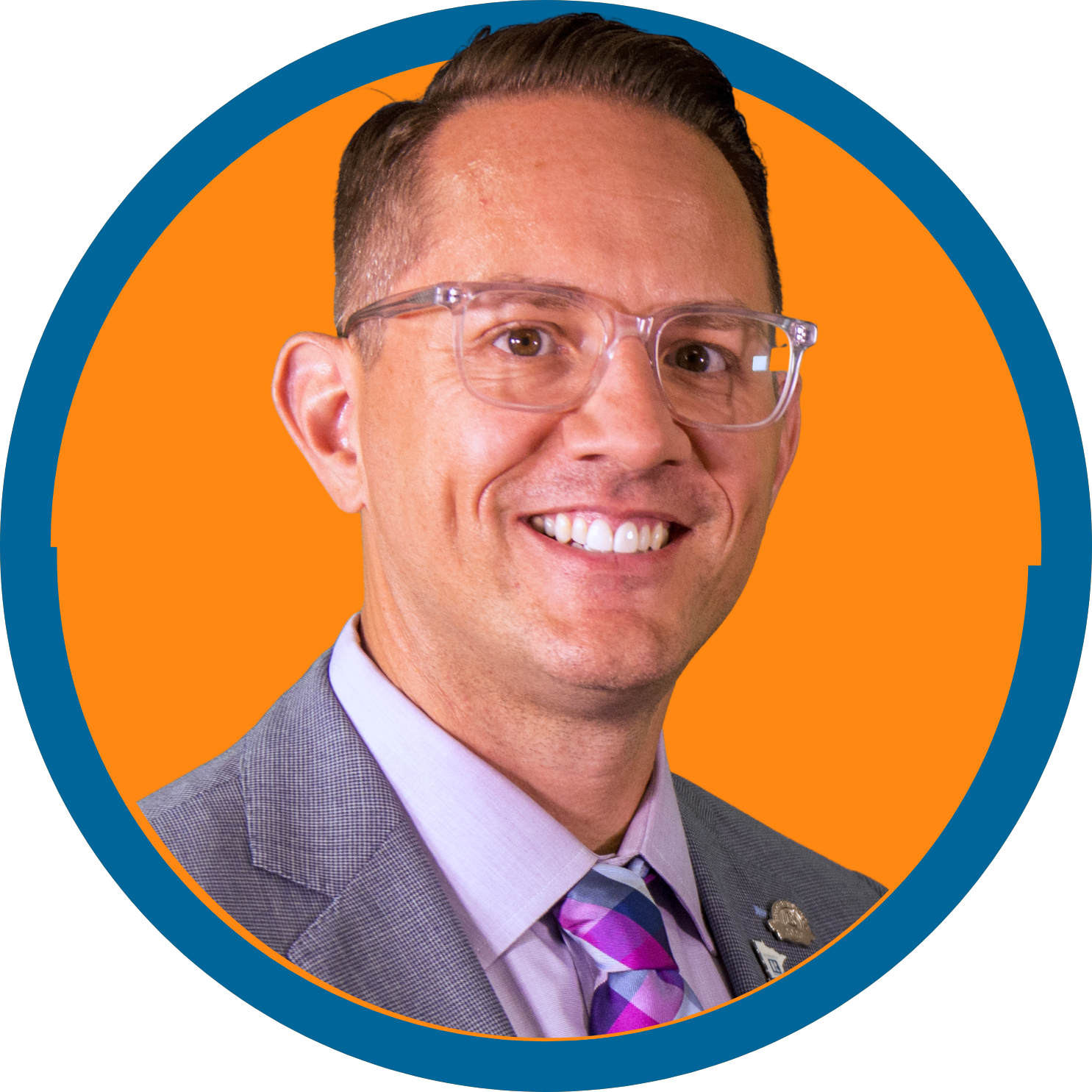 Image of a white man wearing clear glasses, a purple tie and collared shirt, and a gray suit jacket
