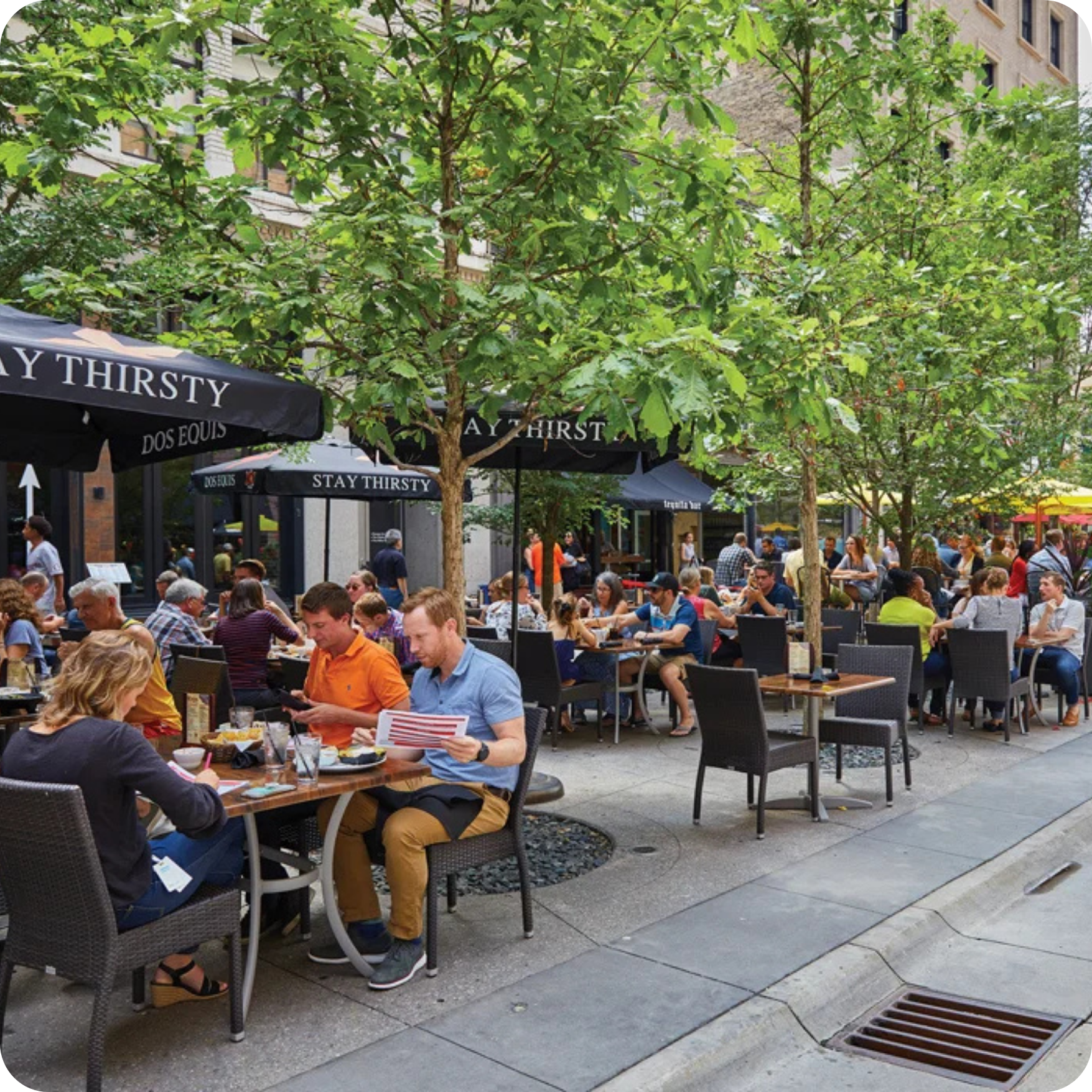 Peopler eating outdoors on a patio with tables, black chairs, trees, and black awnings. 