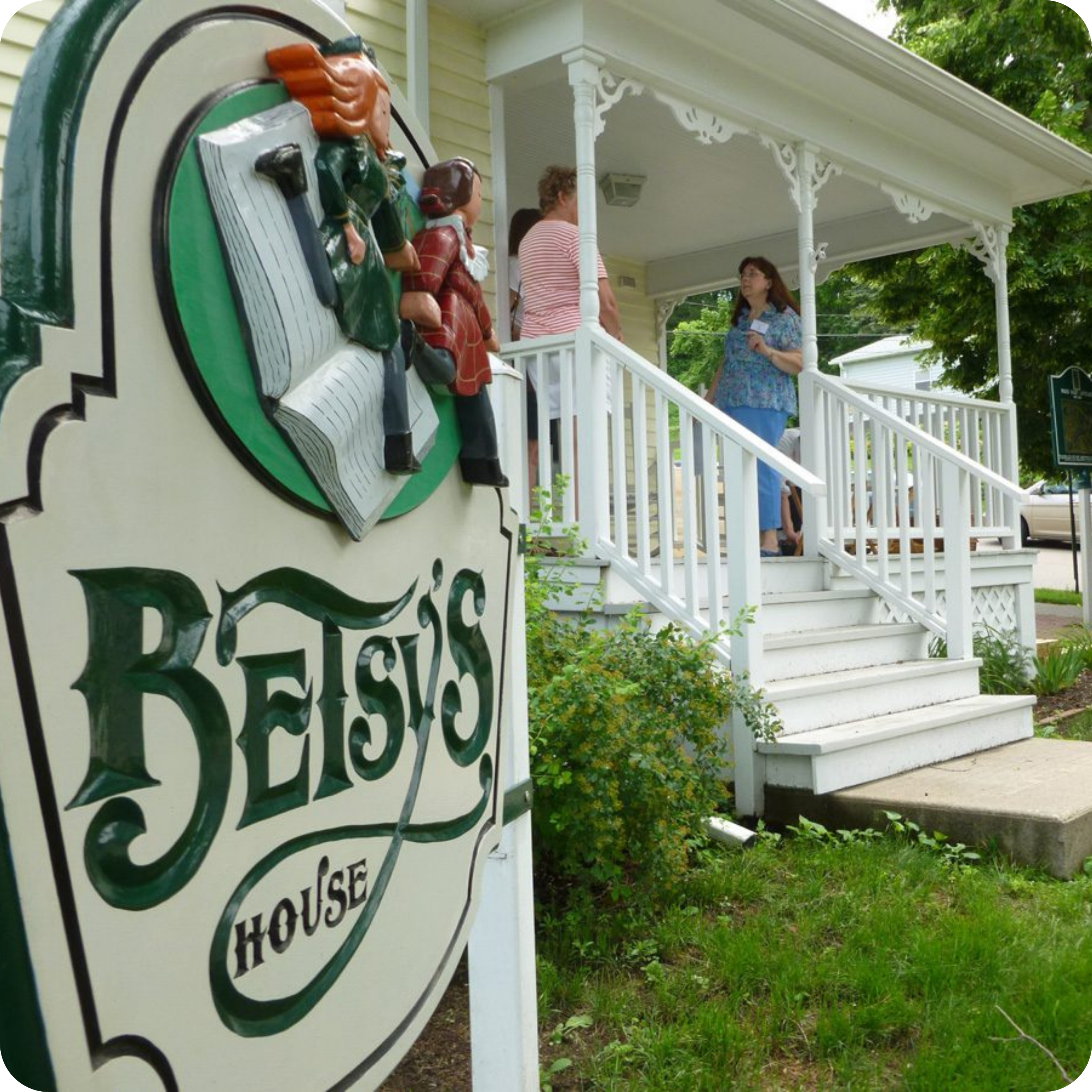 A white building with a decorative porch and three people standing on the steps. In the foreground, a sign reads 