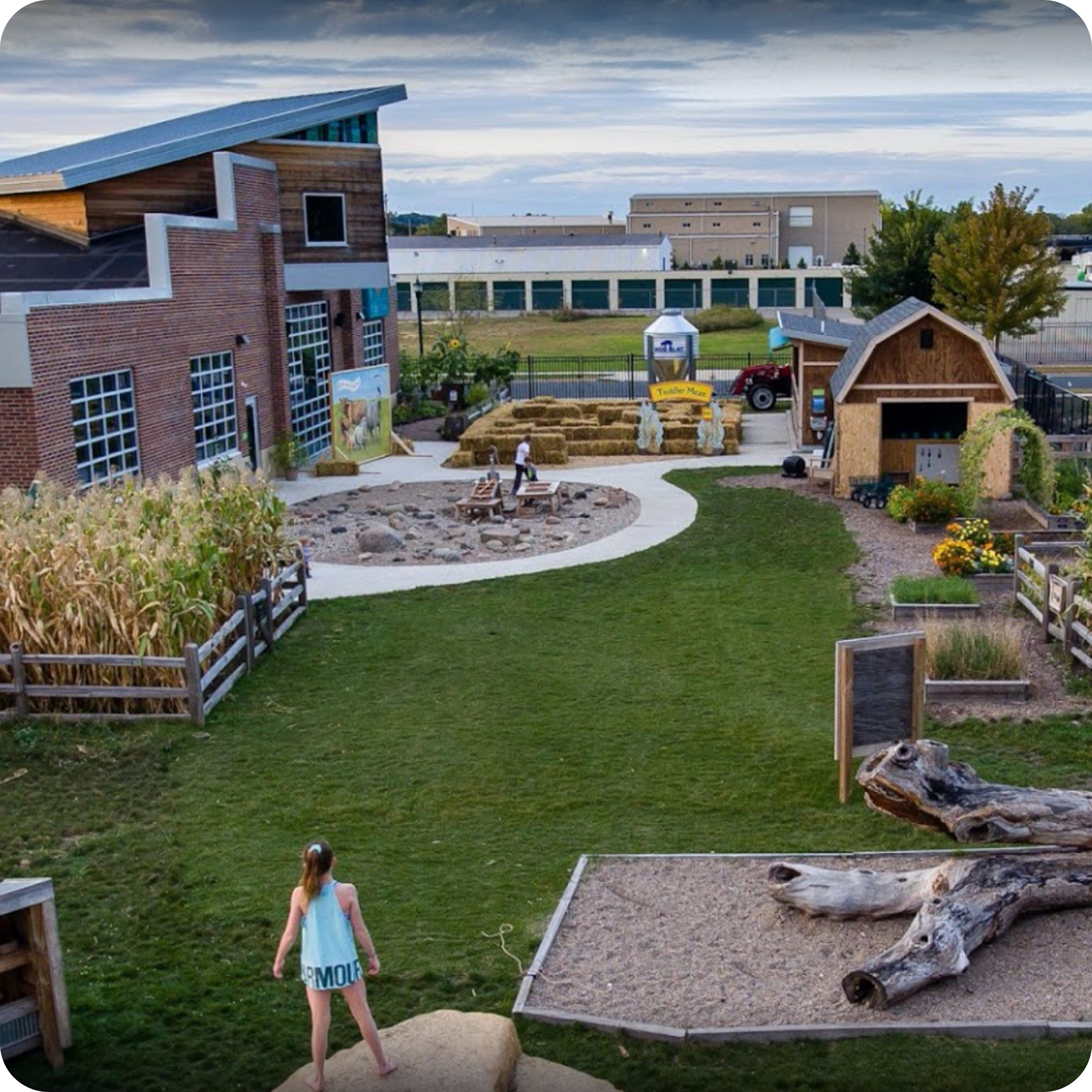 A farm-themed outdoor play area at the Children's Museum of Southern Minnesota, featuring a brick building, corn stalks, and a small barn. The scene includes pathways, logs, hay bales, a tractor, and green spaces, with children playing in various areas.