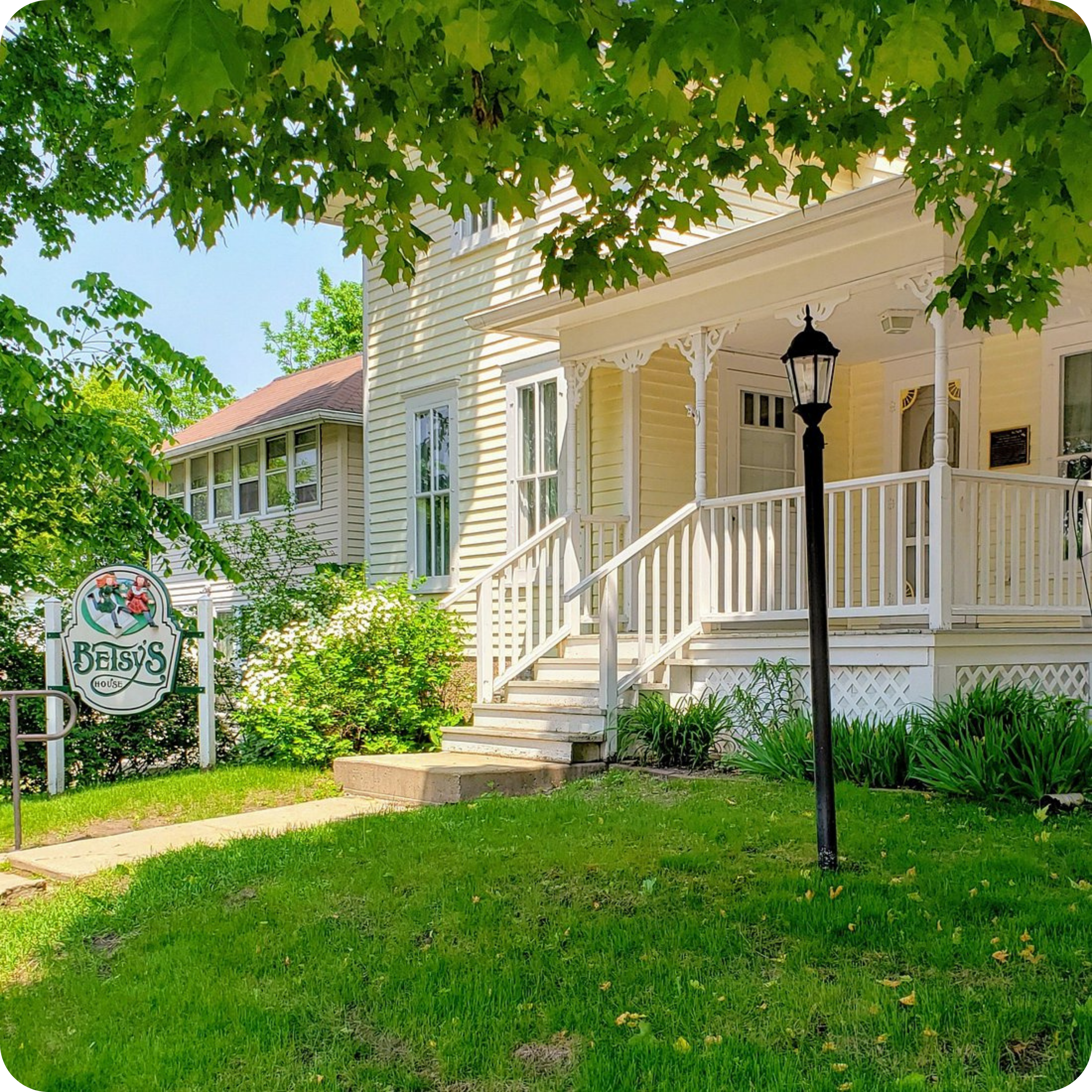 A charming, light yellow house with a welcoming porch, surrounded by green trees and grass. A sign in front displays 