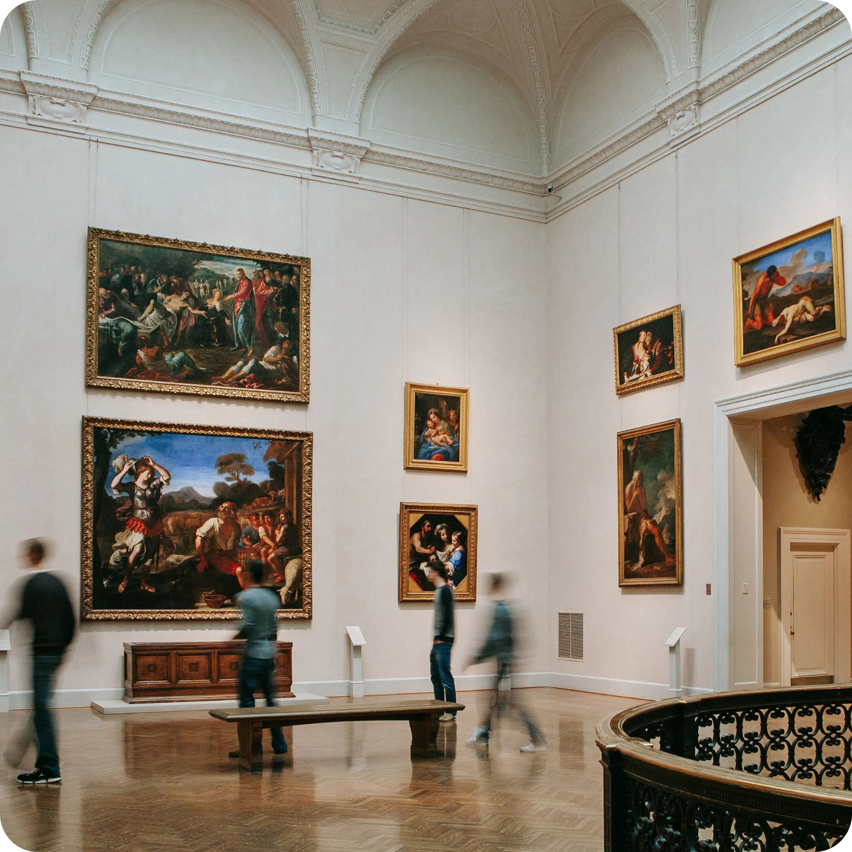 patrons walking through an art gallery with high cream-colored walls and brown flooring.
