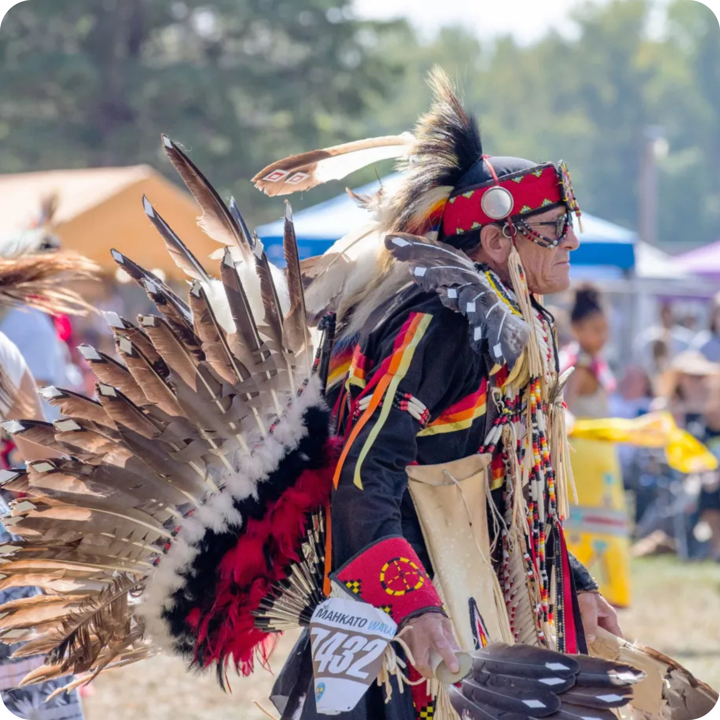 An individual in vibrant traditional regalia participates in a cultural outdoor event. He wears a colorful outfit with feathers and intricate patterns, set against a backdrop of trees and a crowd of onlookers.