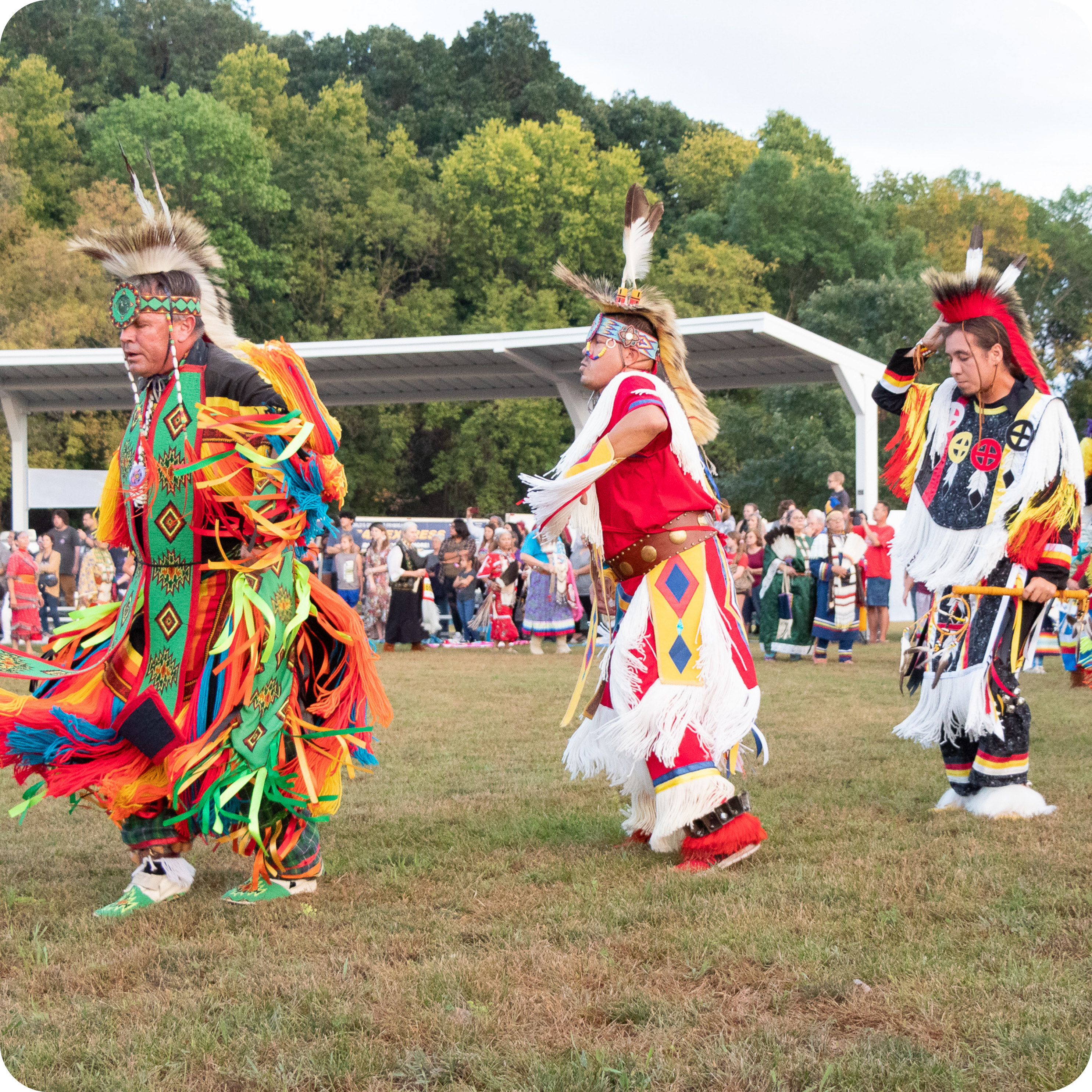 Three individuals in vibrant traditional regalia participate in a cultural dance at an outdoor event. They wear colorful outfits with feathers and intricate patterns, set against a backdrop of trees and a crowd of onlookers.