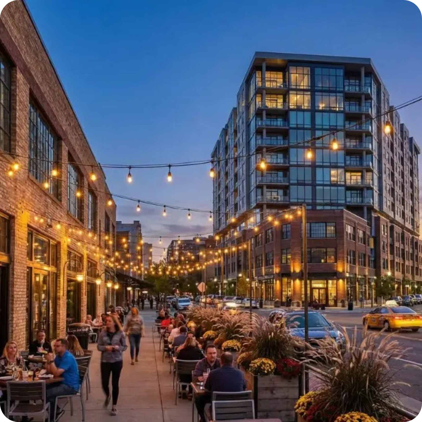 Urban setting at night with an outdoor seating area, lights strung above, and buildings and cars in the background