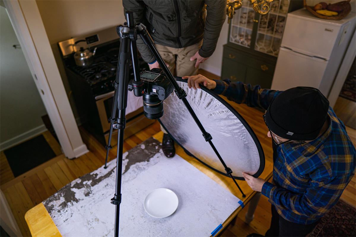 Two people position a light reflector near a camera tripod to make the photo brighter.