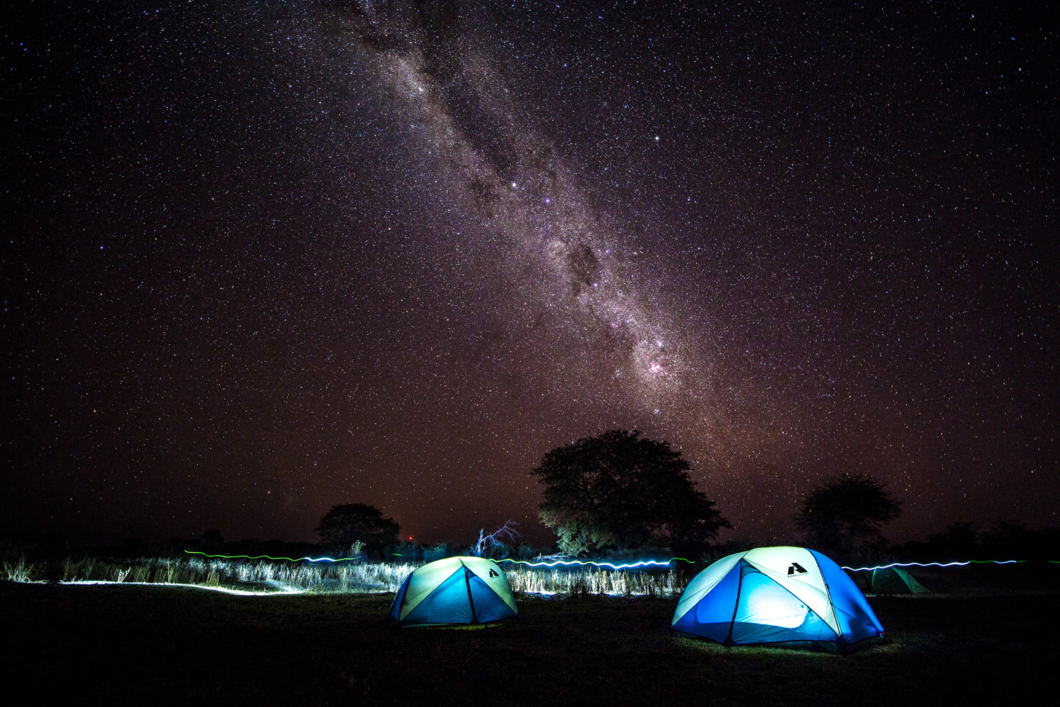 Tents are lit up under a stunning nighttime view of the Milky Way over the Okavango River Delta.