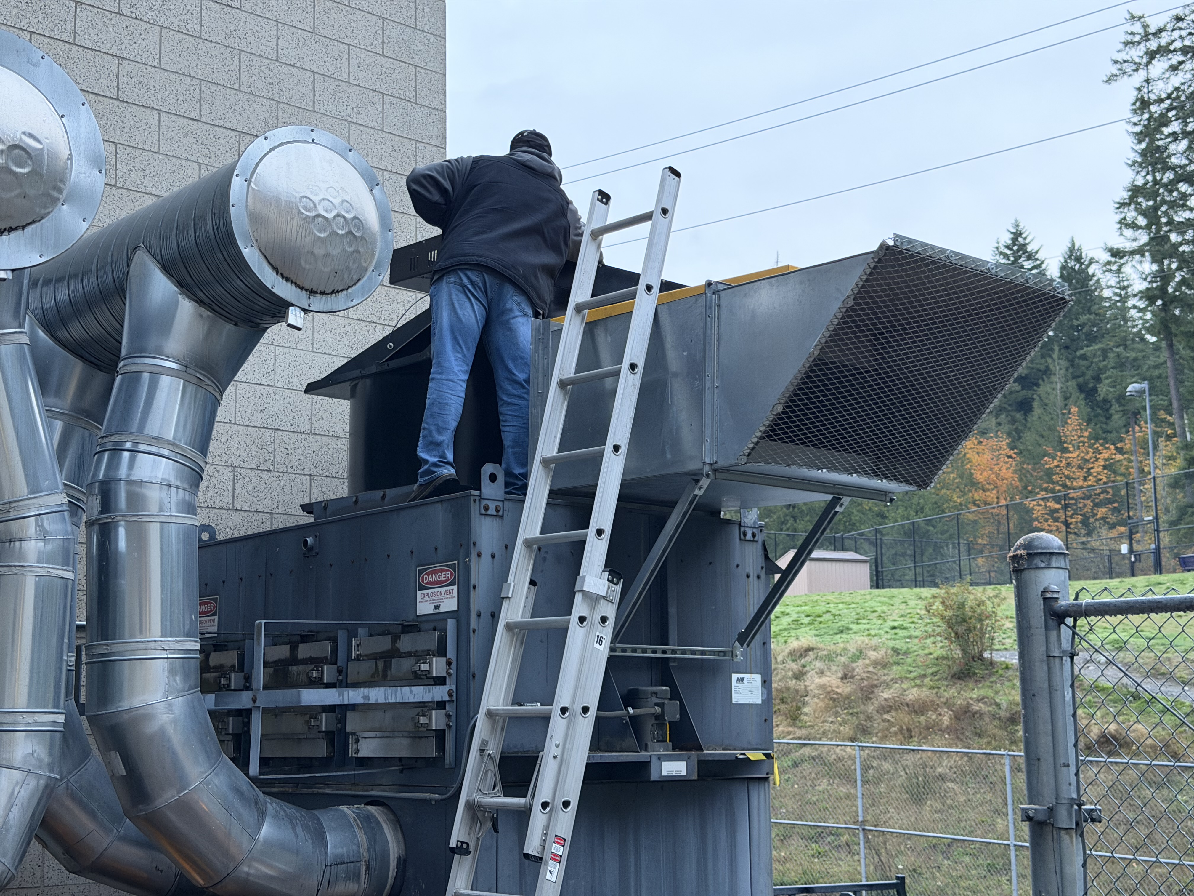 A man on a ladder examines an outdoor piece of equipment used to collect woodshop debrise and keep the air clean 