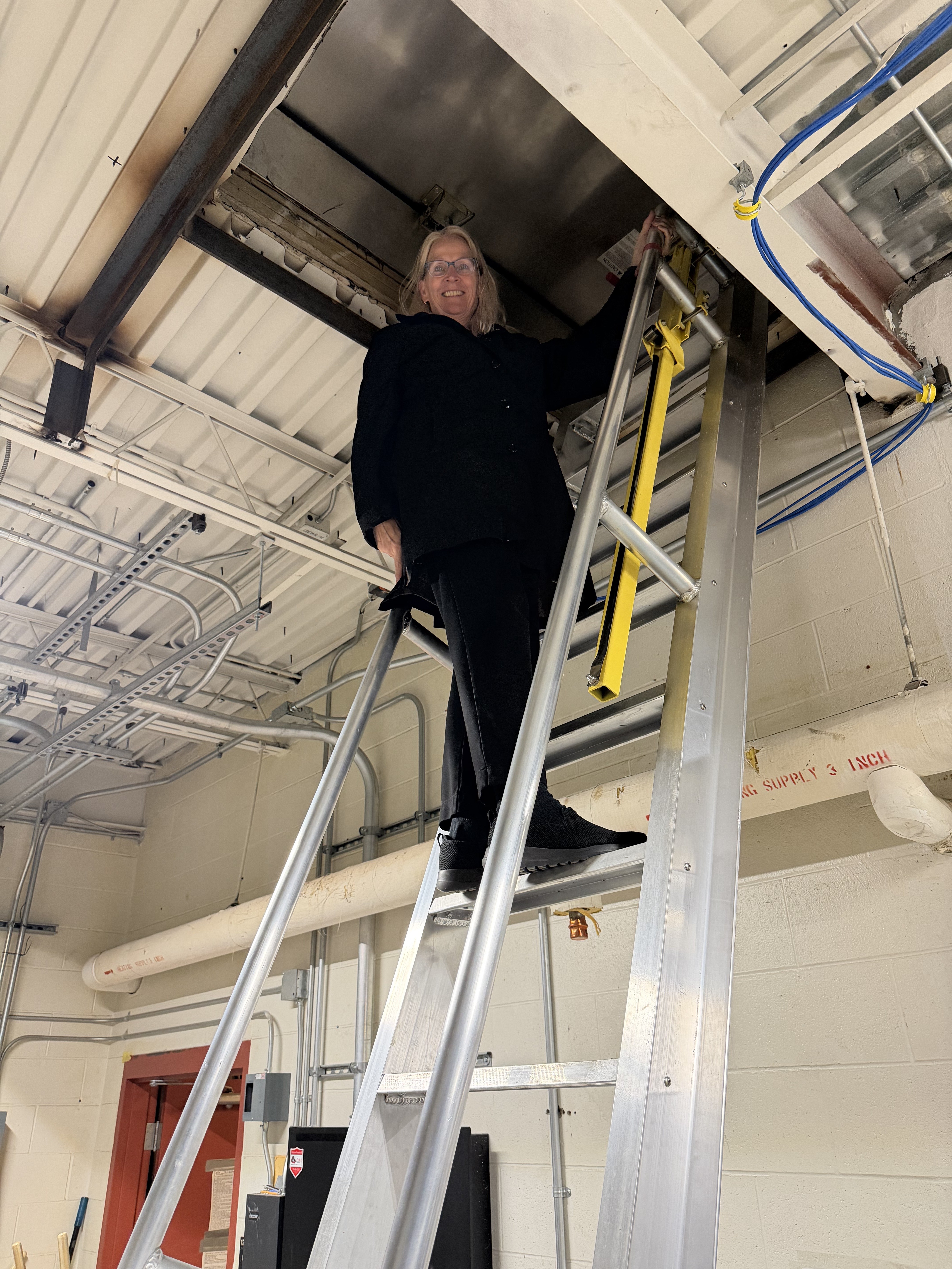 A woman stands on a ladder to access a rooftop HVAC unit. 
