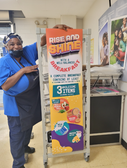 A cafeteria worker stands in front of a sign promoting the school lunch program