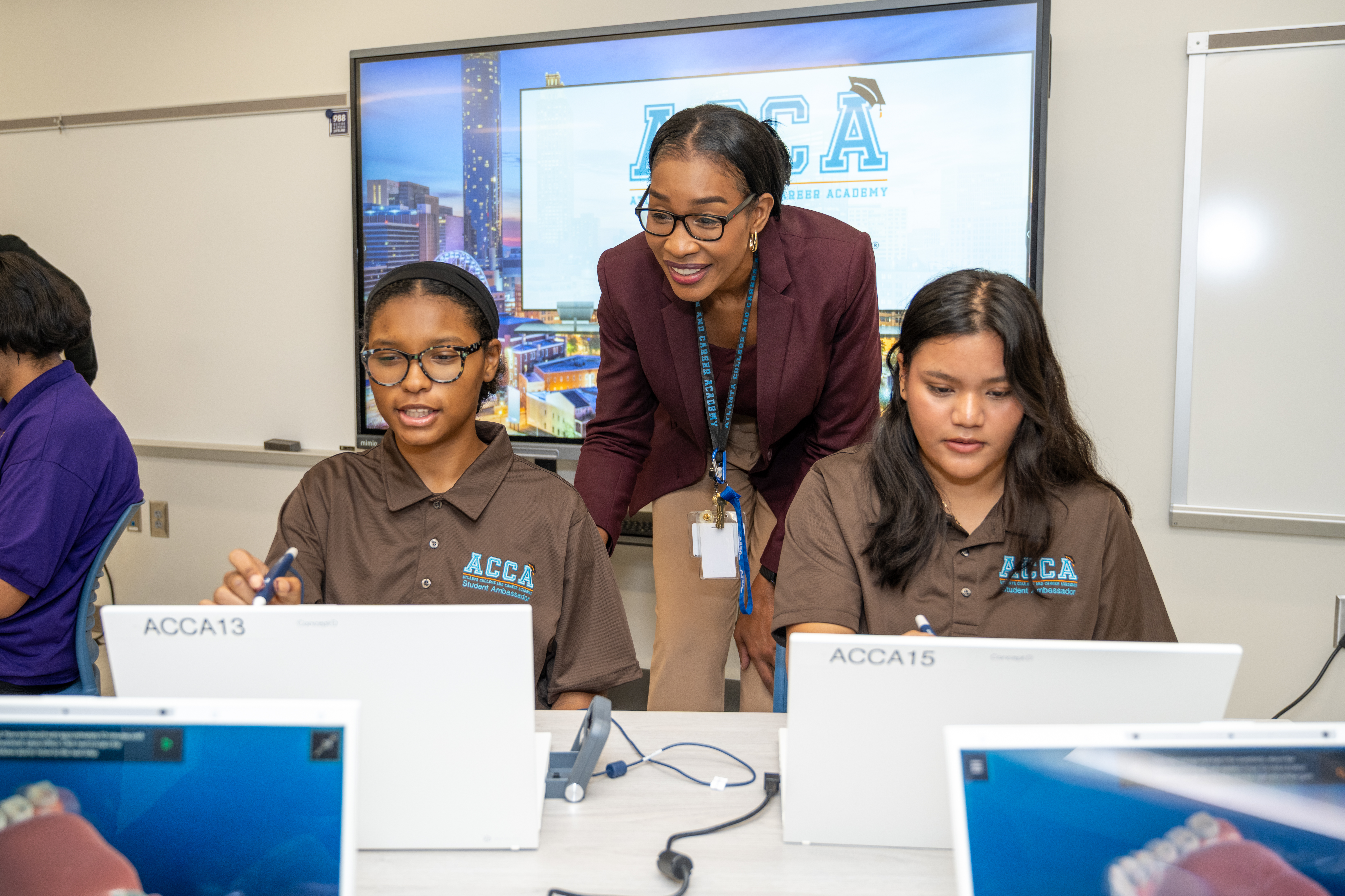 A teacher smiling and looks over the shoulders of two students working on laptops