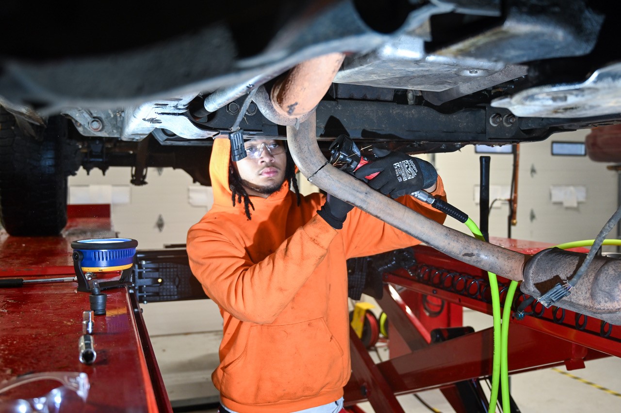 A CAREER AND TECHNOLOGY STUDENT IN AN AUTOMOTIVE PROGRAM PERFORMS REPAIRS UNDER A CAR.
