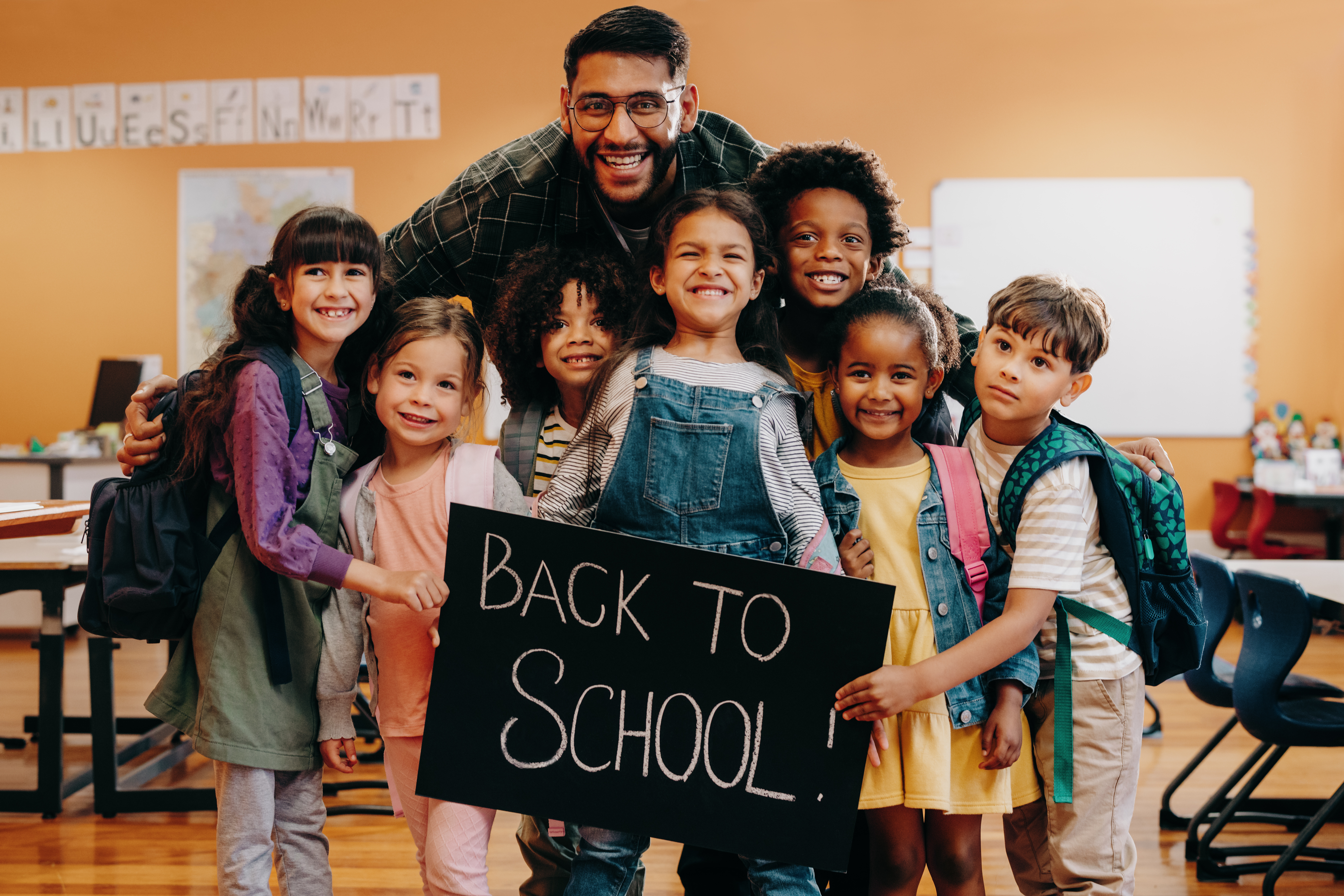 A male teacher smiles with his elementary age students who hold a 'welcome back to school' sign. 