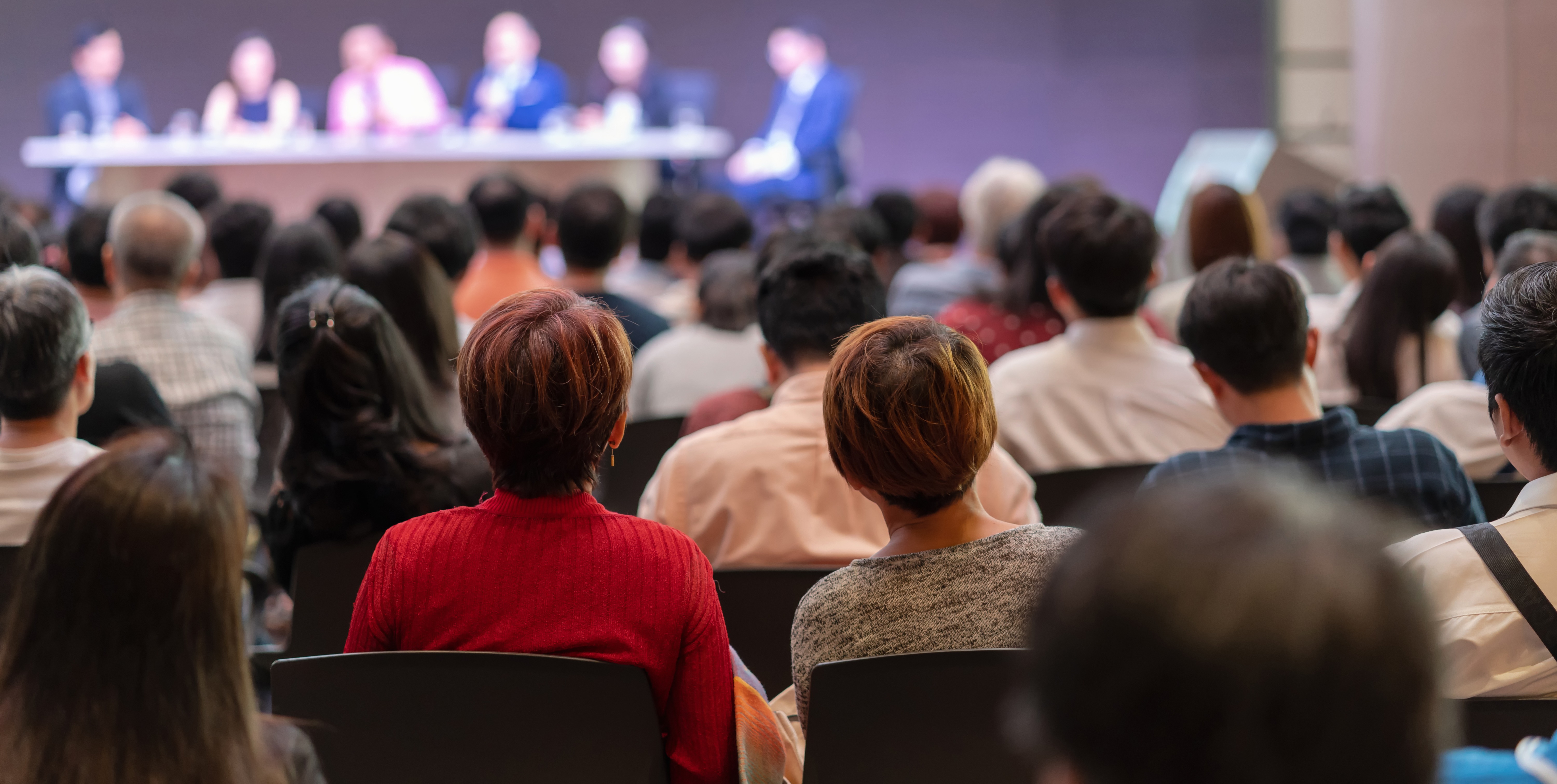 Adults in an auditorium pay attention to a panel discussion on a stage.