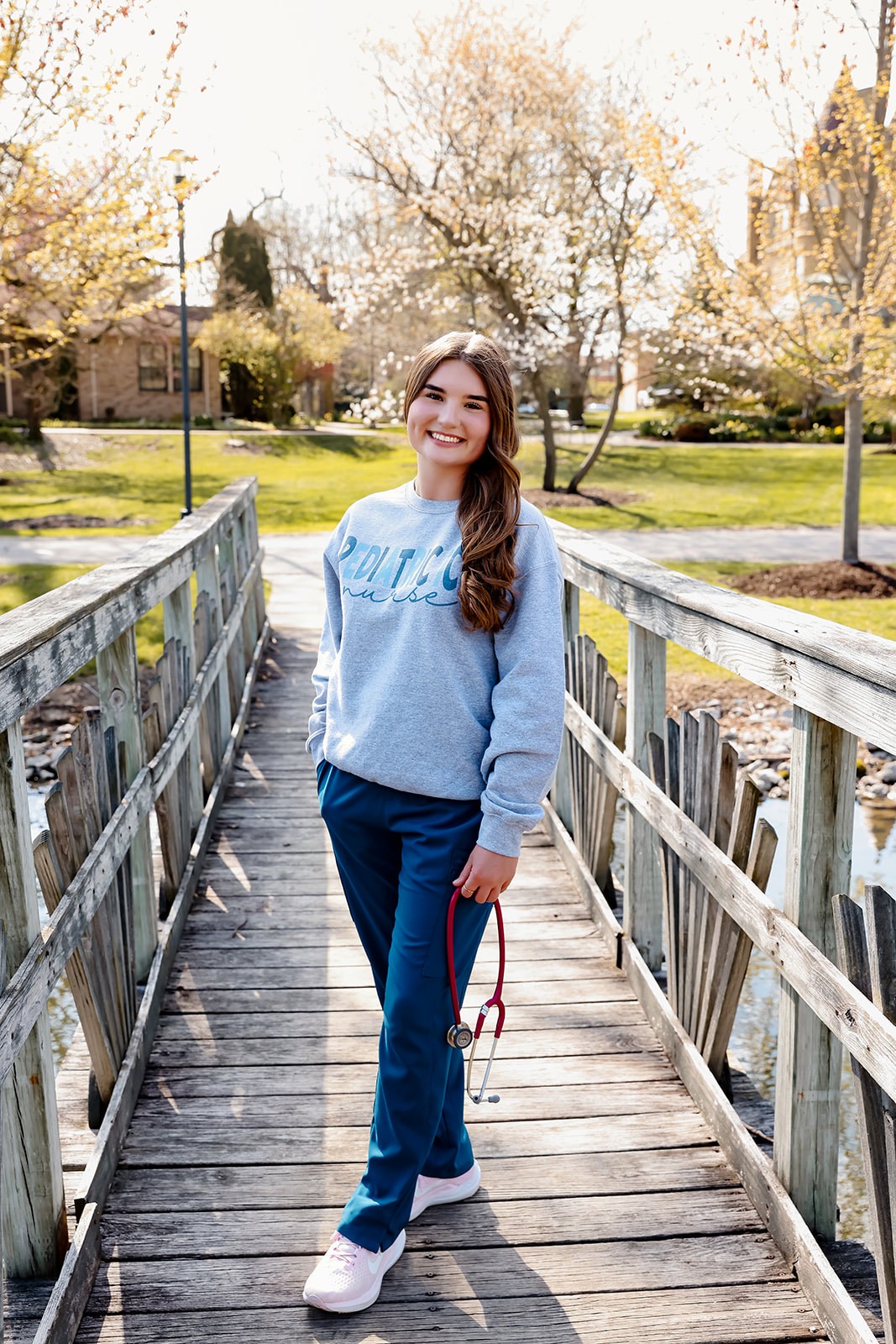 Photo of Lydia Butzin, smiling at the camera. She's standing outside on a sunny day, standing on a wooden bridge and holding a stethoscope. She's wearing a sweatshirt that reads "Pediatric ICU Nurse."