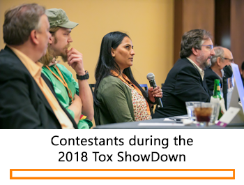 A profile shot of individuals sitting along a long table. The individual in the center of the frame is holding a microphone and looks to be in thought and responding to a question.