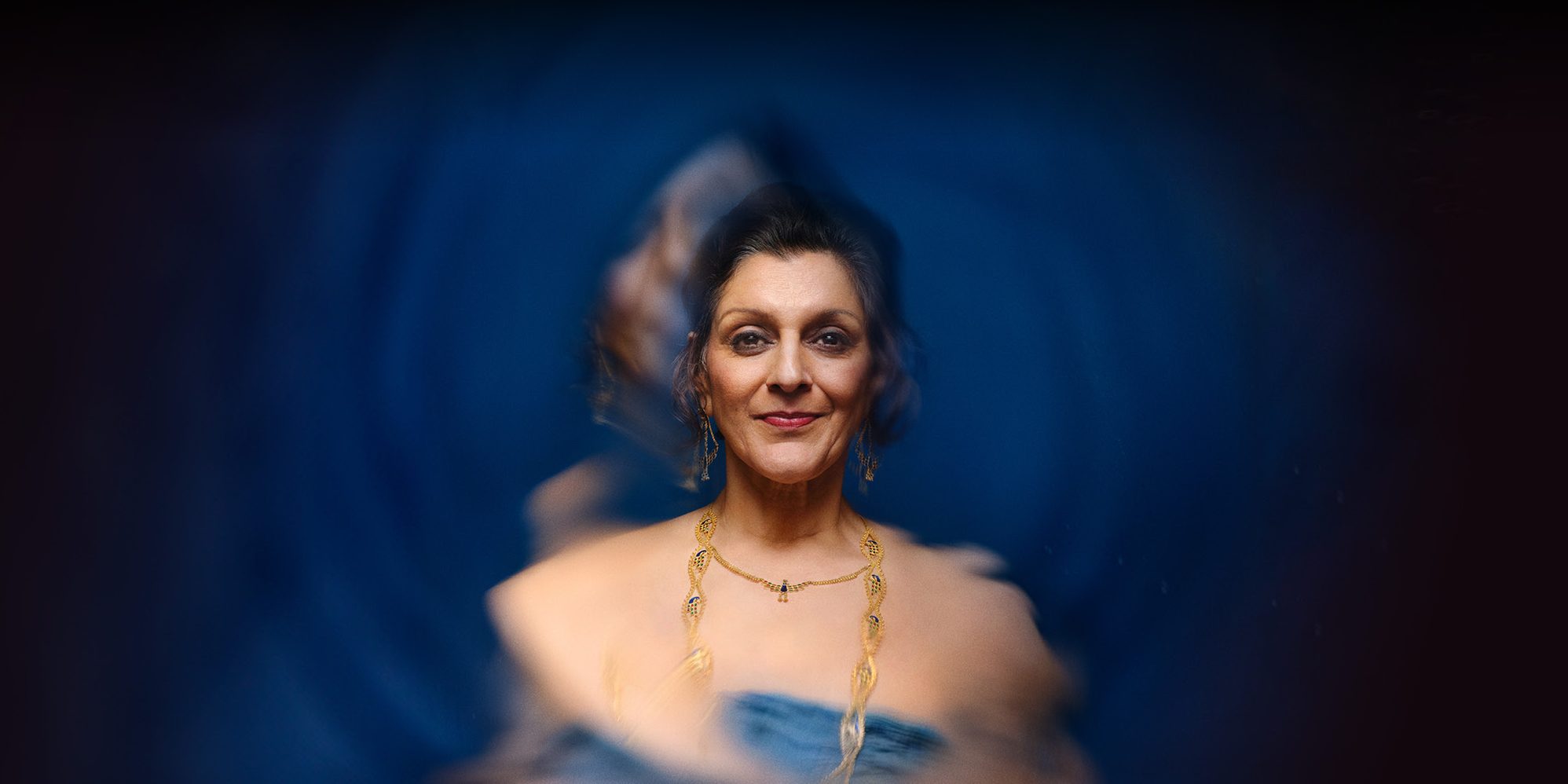 An elegantly dressed South Asian woman in gold jewelry and a blue dressed smiling at the camera