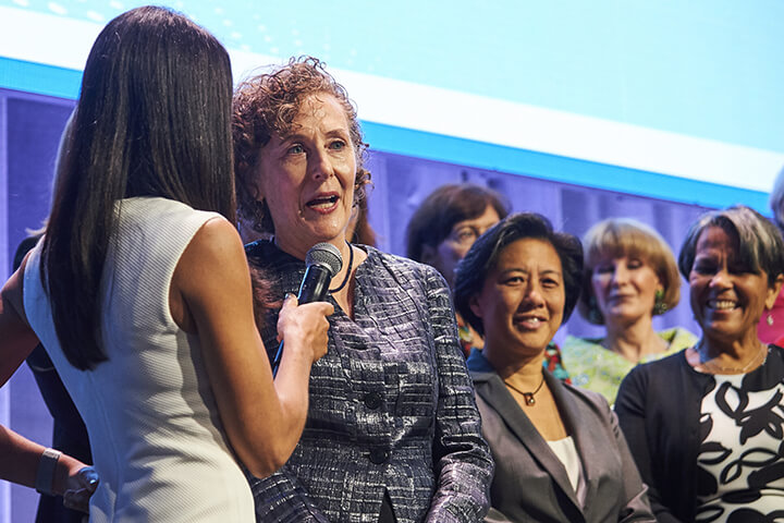 A women speaking into a microphone on stage with other women at the WISE WWOY 2019 Award Luncheon