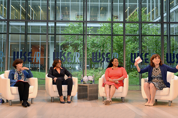 Four women sitting on chairs while the women on the left gesturing as she speaks