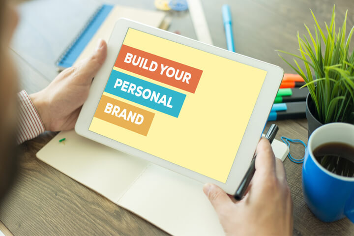 Person holding a tablet with a presentation slide saying "Build your Personal Brand" with plants, a cup of coffee and writing utensils in the background.