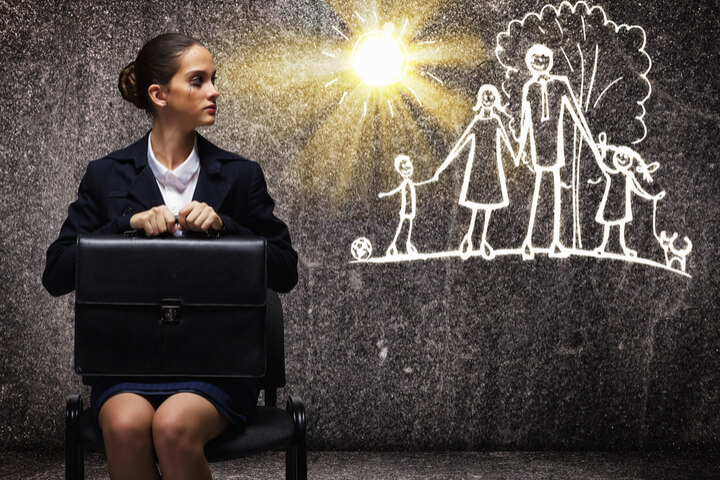 A woman sitting in a chair holding onto a brief case looking sad while looking at an image of a happy family