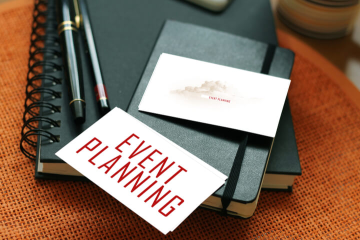 A small notebook layed ontop of another larger notebook, next to pens and a piece of paper saying "event planning"