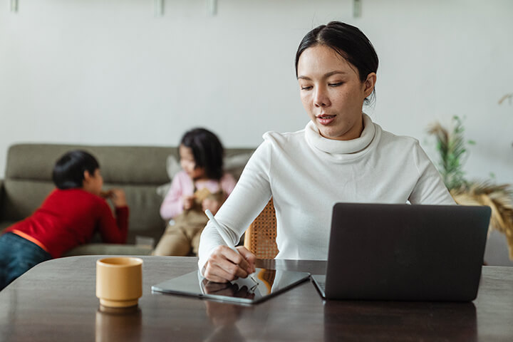 A mother working on her iPad and laptop while children are playing in the background
